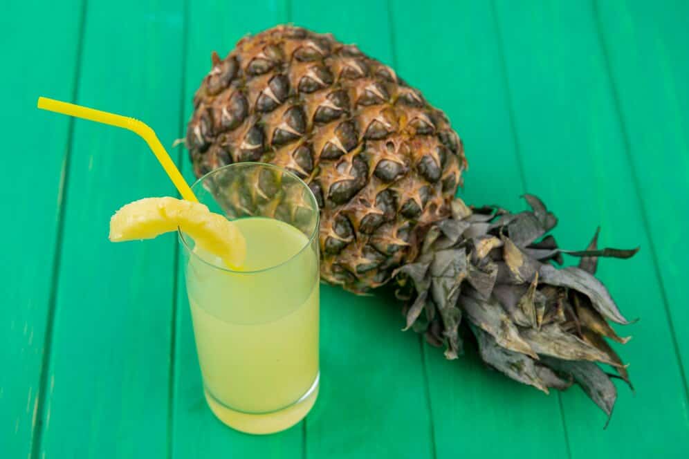 Glass of pineapple and cucumber juice with a pineapple slice and whole pineapple on green wooden surface